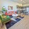 One Bedroom Luxury Apartments in West Sacramento, CA | The Bernice | Living Room with Luxury Vinyl Tile Flooring, Area Rug, Red Sofa, Potted Plant, Standing Lamp, Green Armchair, Round Coffee Table, Two Stools, and View of Kitchen Area.